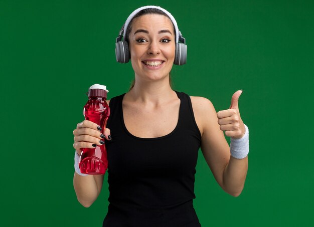 Lächelnde junge hübsche sportliche Frau mit Stirnband und Armbändern mit Kopfhörern, die eine Wasserflasche halten und nach vorne schauen und den Daumen einzeln auf grüner Wand zeigen showing