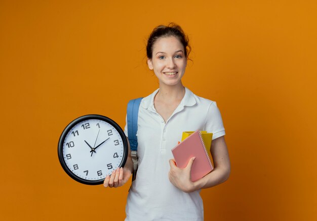 Lächelnde junge hübsche Studentin, die Rückentasche hält Notizblockbuch und Uhr lokalisiert auf orange Hintergrund mit Kopienraum