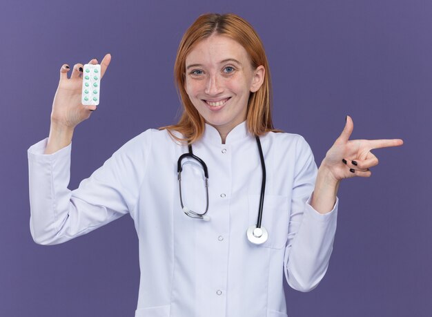 Lächelnde junge Ingwerärztin mit medizinischem Gewand und Stethoskop, die eine Packung medizinischer Pillen auf die Kamera zeigt, die auf die Seite isoliert auf lila Wand zeigt?