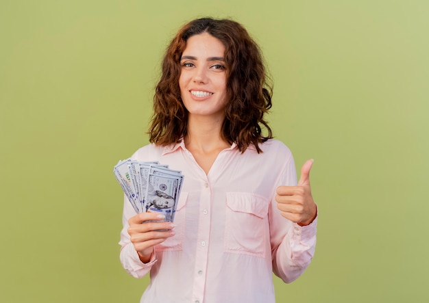 Lächelnde junge kaukasische Frau hält Geld und Daumen lokalisiert auf grünem Hintergrund mit Kopienraum