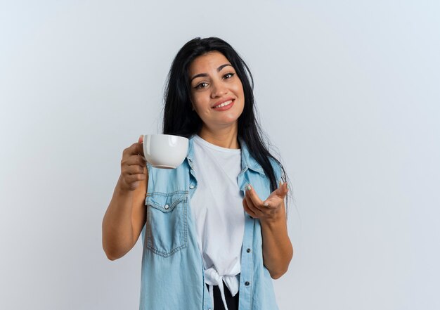 Lächelnde junge kaukasische Frau hält Tasse und zeigt in die Kamera