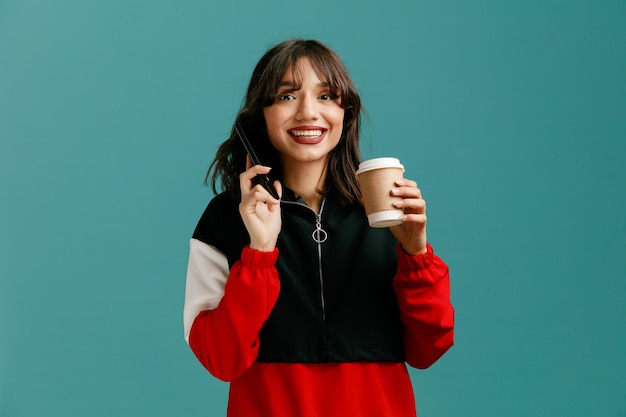 Lächelnde junge kaukasische Frau mit Handy in der Nähe des Ohrs und einer Kaffeetasse zum Mitnehmen in der anderen Hand, die isoliert auf blauem Hintergrund in die Kamera blickt