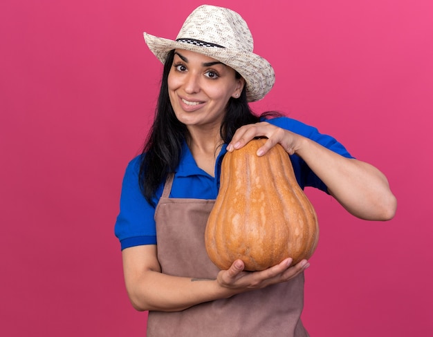 Lächelnde junge kaukasische gärtnerin in uniform und hut mit butternut-kürbis