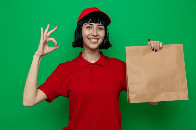 Lächelnde junge kaukasische Lieferfrau, die Papierlebensmittelverpackungen hält und ok Zeichen gestikuliert