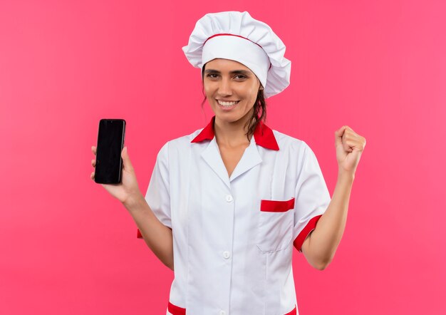 Lächelnde junge Köchin, die Chefuniform hält Telefon hält, zeigt ja Geste mit Kopienraum