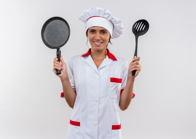 Lächelnde junge Köchin, die Kochuniform trägt, die Bratpfanne und Spatel mit Kopienraum hält