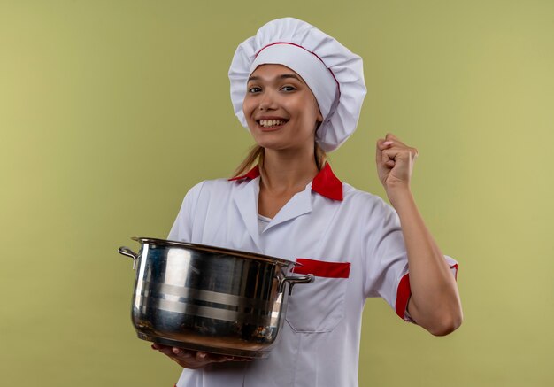 Lächelnde junge Köchin, die Kochuniform trägt, die Topf hält, zeigt ja Geste