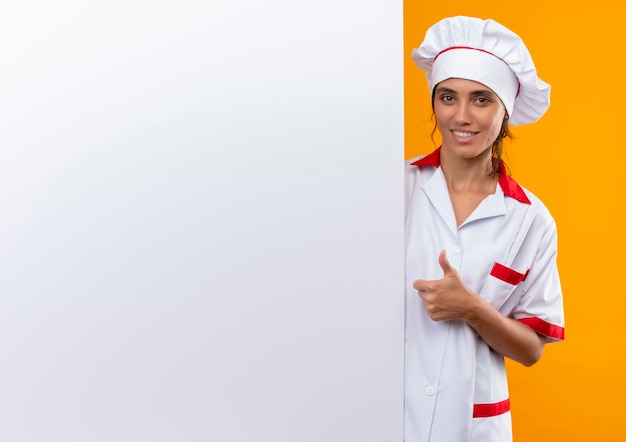 Lächelnde junge Köchin, die Kochuniform trägt, die weiße Wand ihren Daumen oben mit Kopienraum hält