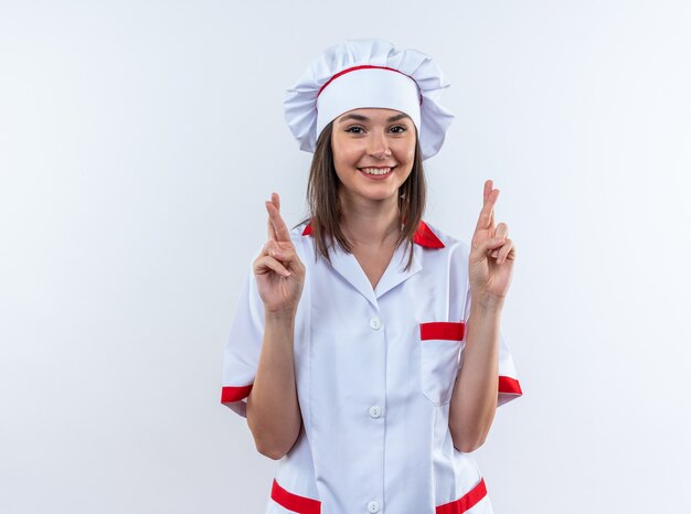 Lächelnde junge Köchin in Kochuniform, die die Finger isoliert auf weißer Wand kreuzt