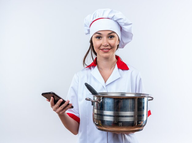 Lächelnde junge Köchin in Kochuniform mit Kochtopf mit Telefon isoliert auf weißer Wand
