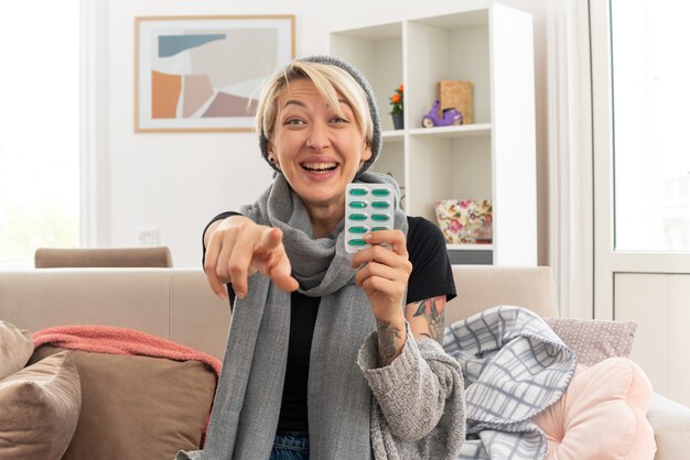 lächelnde junge kranke slawische Frau mit Schal um den Hals, die eine Wintermütze trägt, die eine Medizinblisterpackung hält und nach vorne auf der Couch im Wohnzimmer zeigt