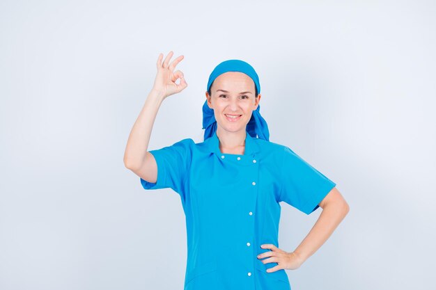 Lächelnde junge Krankenschwester, die eine okay-Geste zeigt und die andere Hand auf die Taille auf weißem Hintergrund legt