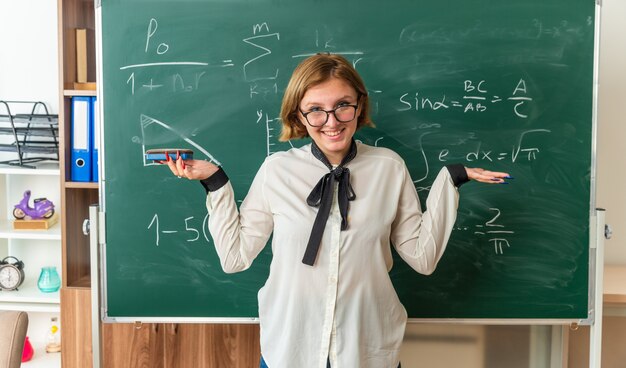 lächelnde junge Lehrerin, die vor Werkzeugen steht und Schwammbrett im Klassenzimmer hält