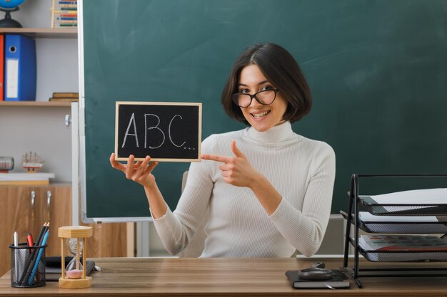 Lächelnde junge Lehrerin mit Brille, die hält und auf eine Mini-Tafel zeigt, die mit Schulwerkzeugen im Klassenzimmer am Schreibtisch sitzt