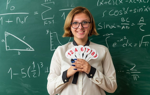 Lächelnde junge lehrerin mit brille, die vor der tafel steht und zahlenfächer im klassenzimmer hält