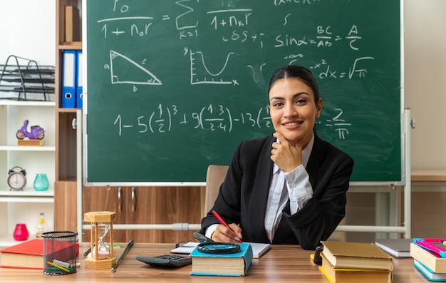 lächelnde junge Lehrerin sitzt am Tisch mit Schulmaterial und schreibt etwas am Kinn gepackt im Klassenzimmer