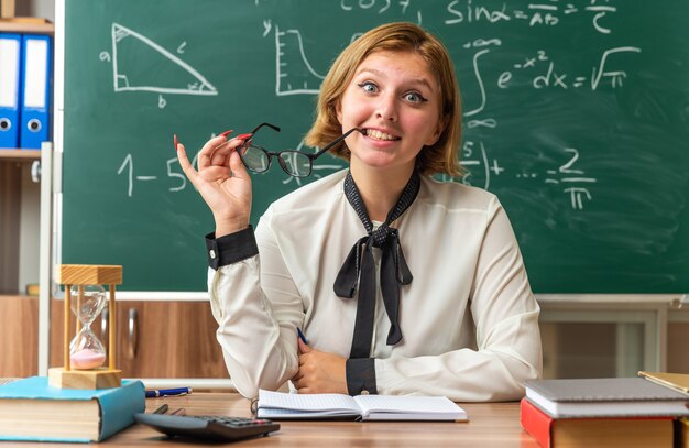 Lächelnde junge Lehrerin sitzt am Tisch mit Schulwerkzeugen und hält eine Brille im Klassenzimmer holding