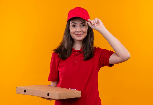 Lächelnde junge Lieferfrau, die rotes T-Shirt in der roten Kappe hält, die eine Pizzaschachtel auf isolierter orange Wand hält
