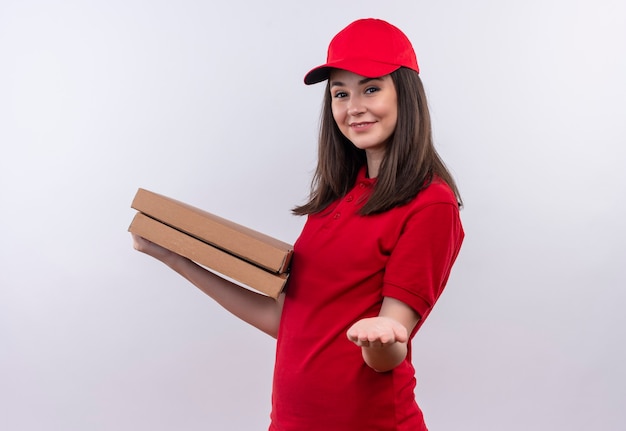Lächelnde junge Lieferfrau, die rotes T-Shirt in der roten Kappe hält, die eine Pizzaschachtel hält und ihre Hand auf isolierter weißer Wand aushält