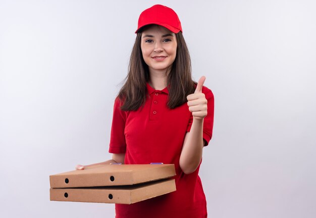 Lächelnde junge Lieferfrau, die rotes T-Shirt in der roten Kappe hält, die Pizzaschachtel hält und Daumen oben auf isolierter weißer Wand zeigt