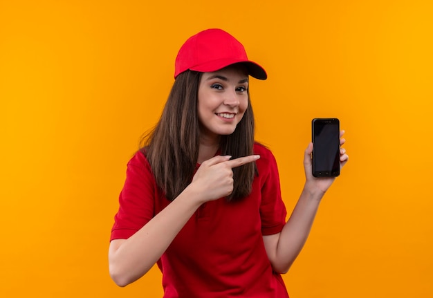 Lächelnde junge Lieferfrau, die rotes T-Shirt in der roten Kappe hält Telefon auf der einen Hand hält und mit der anderen Hand auf isolierte gelbe Wand darauf zeigt