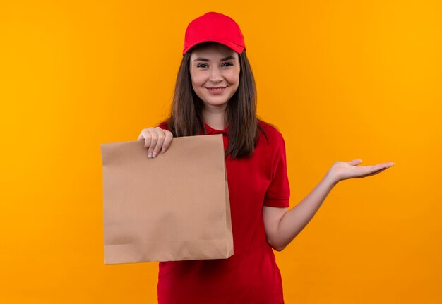 Lächelnde junge Lieferfrau, die rotes T-Shirt in der roten Kappe trägt, die eine Tasche auf isolierter orange Wand hält