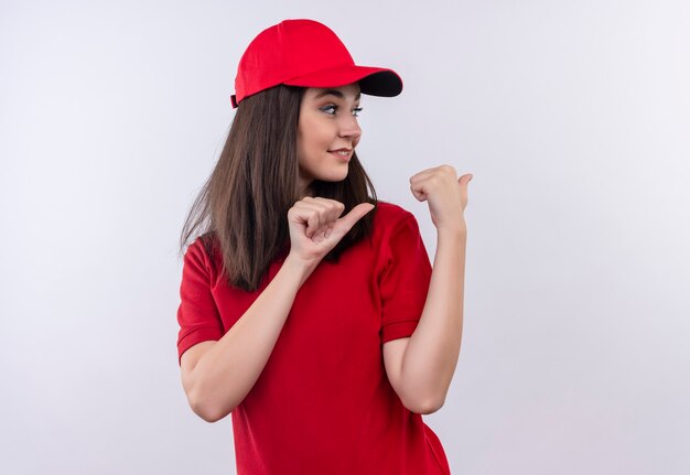 Lächelnde junge Lieferfrau, die rotes T-Shirt in der roten Kappe trägt, zeigt auf isolierte weiße Wand zurück