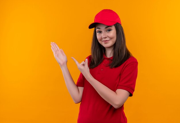 Lächelnde junge Lieferfrau, die rotes T-Shirt in roter Kappe trägt und Hand und Finger zur Seite auf isolierte orange Wand zeigt