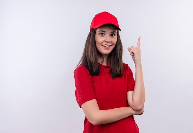 Lächelnde junge Lieferfrau, die rotes T-Shirt in roter Kappe trägt, zeigt auf oben auf isolierter weißer Wand