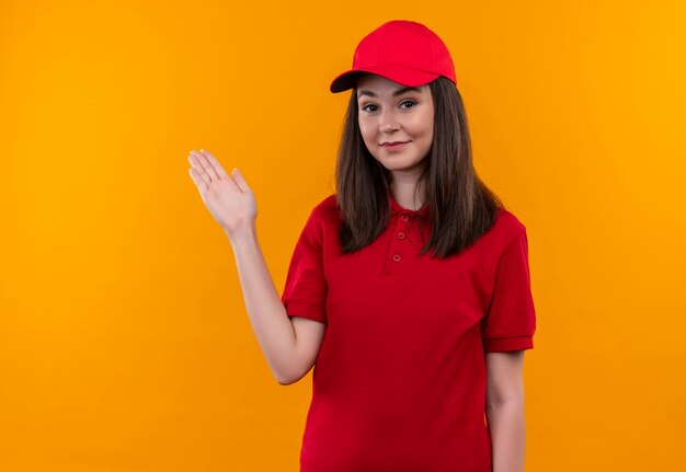 Lächelnde junge Lieferfrau, die rotes T-Shirt in roter Kappe trägt, zeigt zur Seite mit ihrer Hand auf isolierter orange Wand