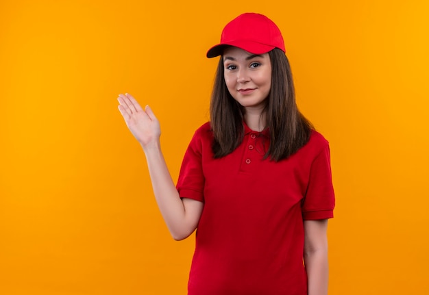 Lächelnde junge Lieferfrau, die rotes T-Shirt in roter Kappe trägt, zeigt zur Seite mit ihrer Hand auf isolierter orange Wand