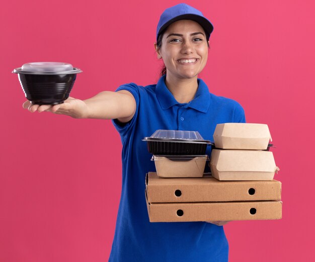 Lächelnde junge Lieferfrau, die Uniform mit Kappe trägt, die Lebensmittelbehälter auf Pizzakästen hält, die Lebensmittelbehälter an der Kamera lokalisiert auf rosa Wand halten