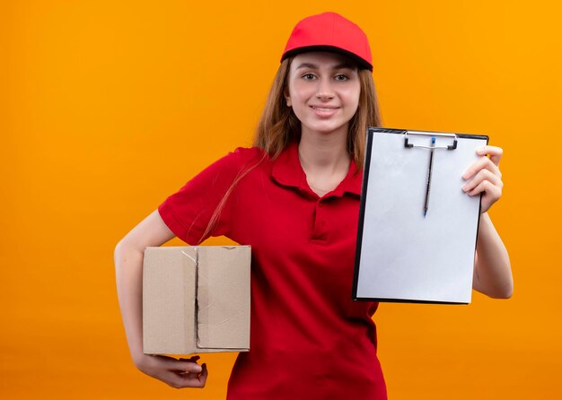 Lächelnde junge Lieferfrau in der roten Uniform, die Box hält und Zwischenablage auf lokalisiertem orange Raum zeigt