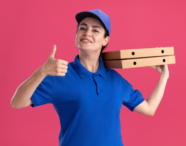 Lächelnde junge Lieferfrau in Uniform und Mütze, die Pizzapakete auf der Schulter hält und den Daumen zeigt