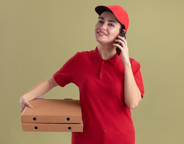 Lächelnde junge lieferfrau in uniform und mütze mit pizzapaketen, die am telefon telefonieren