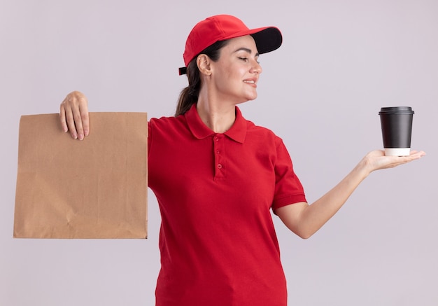 Lächelnde junge Lieferfrau in Uniform und Mütze mit Plastikkaffeetasse und Papierpaket mit Blick auf die Kaffeetasse isoliert auf weißer Wand