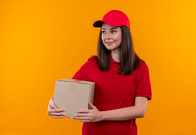 Lächelnde junge Lieferung lächelnde Frau im roten T-Shirt und in der roten Kappe, die eine Box auf orange Wand hält