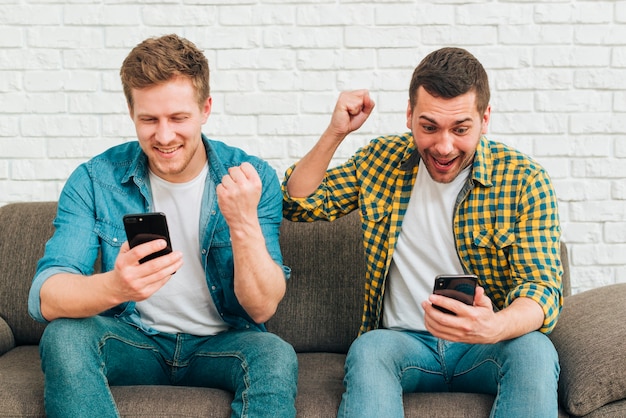 Lächelnde junge männliche Freunde, die das intelligente Telefon zusammenpreßt ihre Faust betrachten