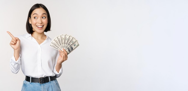 Lächelnde junge moderne asiatische frau, die auf die bannerwerbung zeigt, die bargelddollar hält, die über weißem hintergrund stehen
