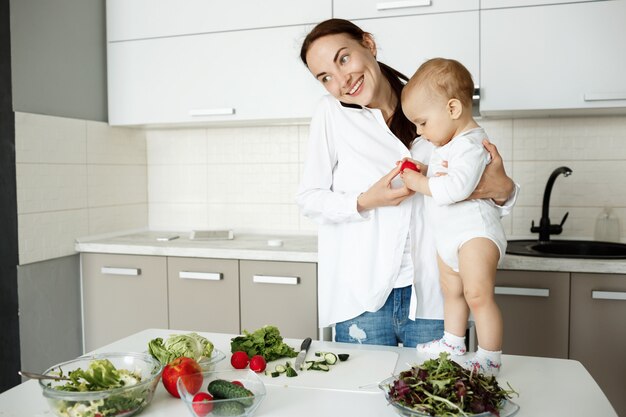 Lächelnde junge Mutter, die Baby hält, am Telefon spricht und gesundes Frühstück vorbereitet