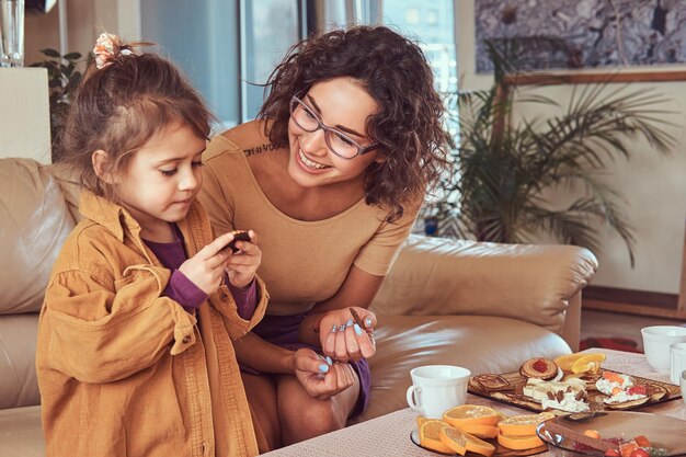 Lächelnde junge Mutter mit süßer kleiner Tochter, die zu Hause auf dem Sofa frühstückt.