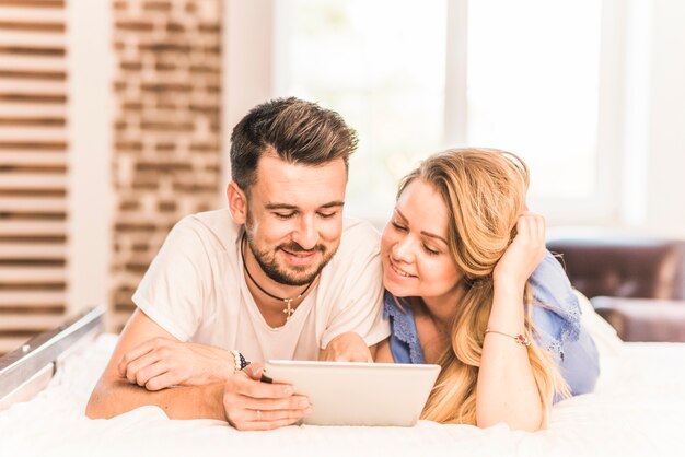 Lächelnde junge Paare, die auf Bett unter Verwendung der digitalen Tablette liegen