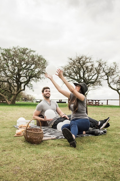 Lächelnde junge Paare, die mit den weißen Ballonen sitzen im Park spielen