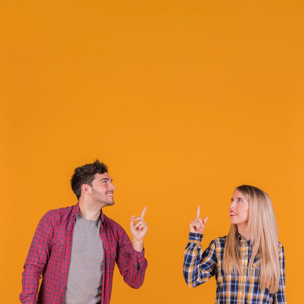 Lächelnde junge Paare, die oben schauen und aufwärts ihren Finger gegen einen orange Hintergrund zeigen