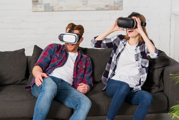 Lächelnde junge Paare, die zu Hause auf schwarzem Sofa unter Verwendung eines vr Kopfhörers sitzen