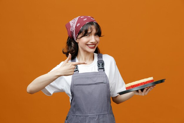 Lächelnde junge Putzfrau in Uniform und Bandana, die ein Tablett mit einem Schwamm darin hält und auf das Tablett zeigt, das isoliert auf orangefarbenem Hintergrund in die Kamera blickt