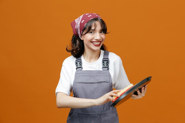 Lächelnde junge Putzfrau in Uniform und Kopftuch mit Blick auf die Kamera-Reinigungsschale mit Schwamm isoliert auf orangefarbenem Hintergrund