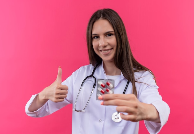 Lächelnde junge Ärztin, die das medizinische Kleid des Stethoskops trägt, hält Pillen ihren Daumen auf lokalisiertem rosa Hintergrund