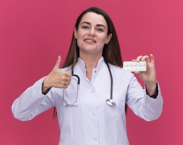 Lächelnde junge Ärztin, die ein medizinisches Gewand mit Stethoskop trägt, hält eine Packung Medizin und Daumen hoch isoliert auf rosa Wand mit Kopierraum