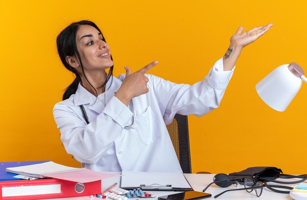 Lächelnde junge Ärztin, die ein medizinisches Gewand mit Stethoskop trägt, sitzt am Tisch mit medizinischen Instrumenten, die vorgeben, zu halten, und zeigt auf etwas, das auf gelbem Hintergrund isoliert ist